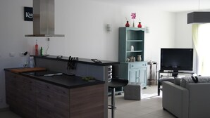 A modern kitchen with a central island, a dining area with a grey sofa, a TV, and a teal cabinet with decorative items. - Family home 1000m from the ocean (Vensac)