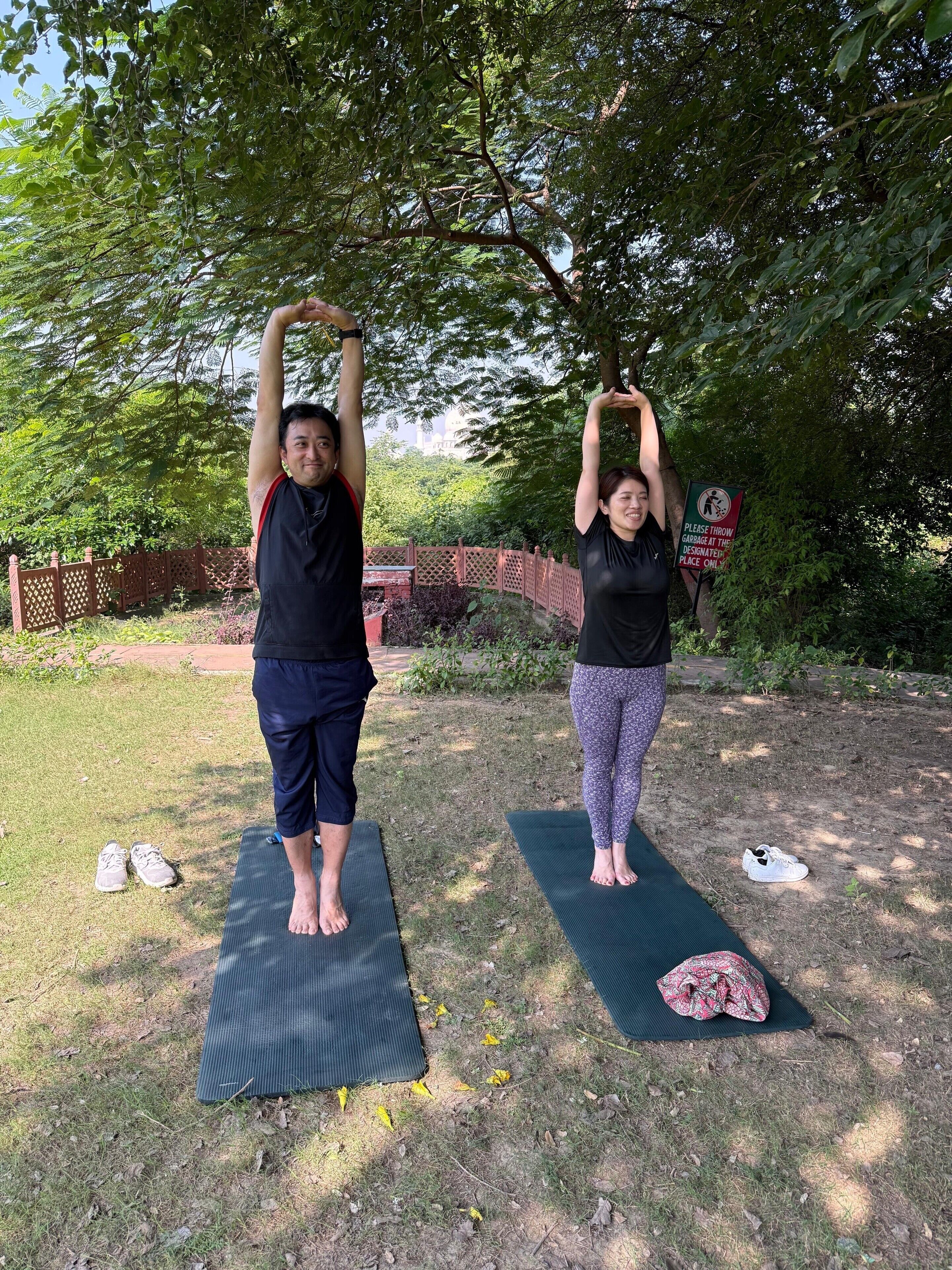 Zwei Personen üben Yoga auf Matten im Freien, im Hintergrund Bäume und ein Zaun.