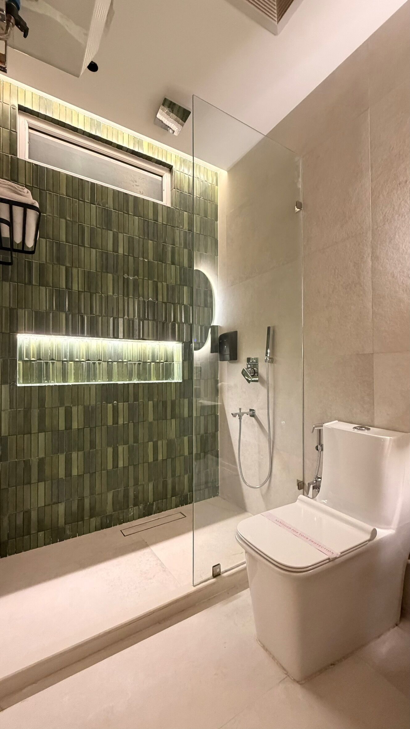 Un baño moderno con ducha envuelta en vidrio, inodoro blanco y pared con azulejos verdes e iluminación.