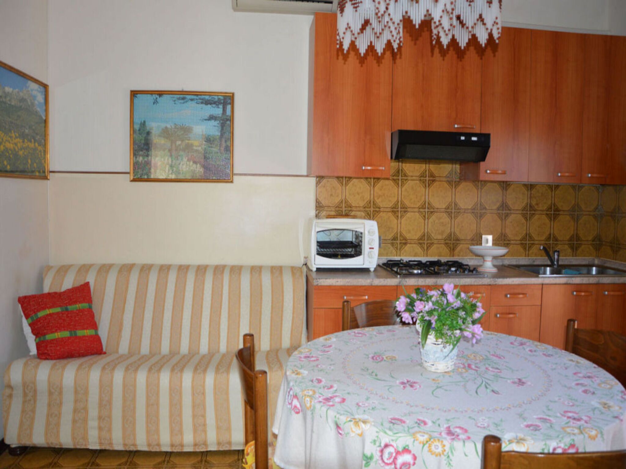A cozy living room with a striped sofa, a small table with a floral tablecloth, and a kitchen area with a toaster and stove.