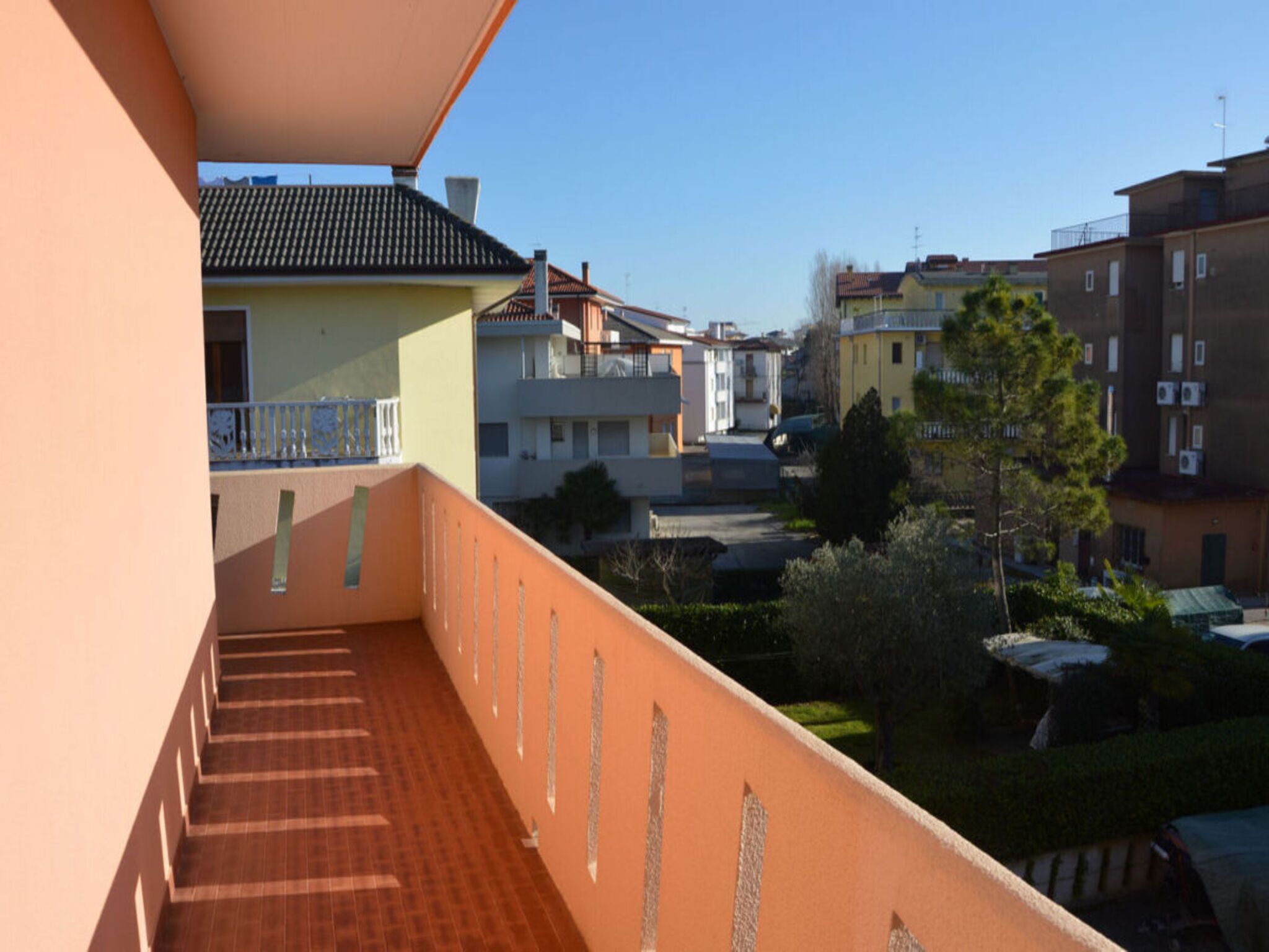 A balcony with a tiled floor and a railing, overlooking a residential area with various buildings and a clear sky.