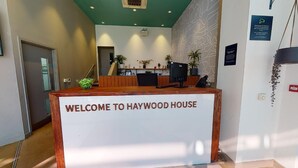 Un comptoir d’accueil avec une pancarte indiquant « BIENVENUE À HAYWOOD HOUSE », une télévision et des plantes en pot.