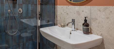 Un baño moderno con ducha envuelta en vidrio, un lavamanos blanco con dispensador de jabón y un espejo redondo.
