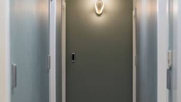 A narrow hallway with a light fixture on the wall, a door at the end, and a wooden floor.