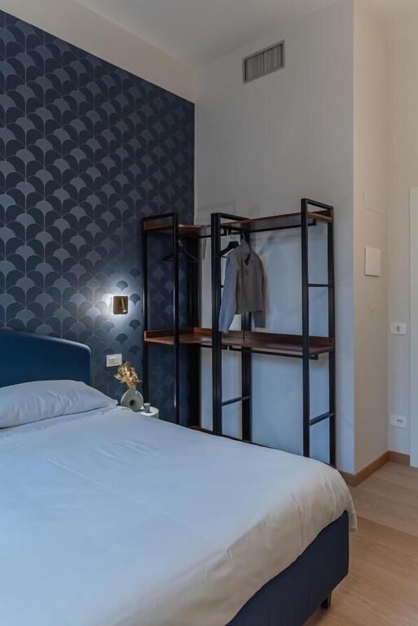 A bedroom with a bed, a wall-mounted shelf with clothes, and a small table with a vase. - San Gregorio Rooms Milano Centrale (Milan)
