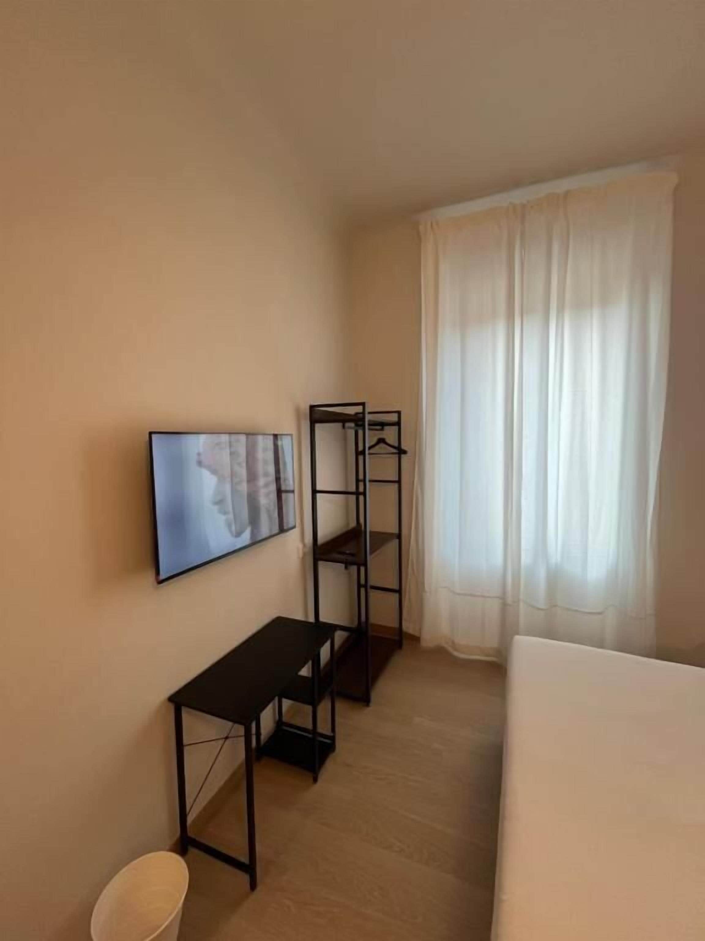 A hotel room with a bed, a TV mounted on the wall, a small table, a coat rack, and a window with curtains.
