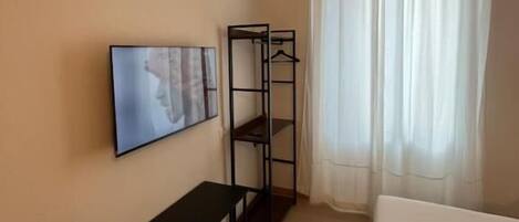 A hotel room with a bed, a TV mounted on the wall, a small table, a coat rack, and a window with curtains.