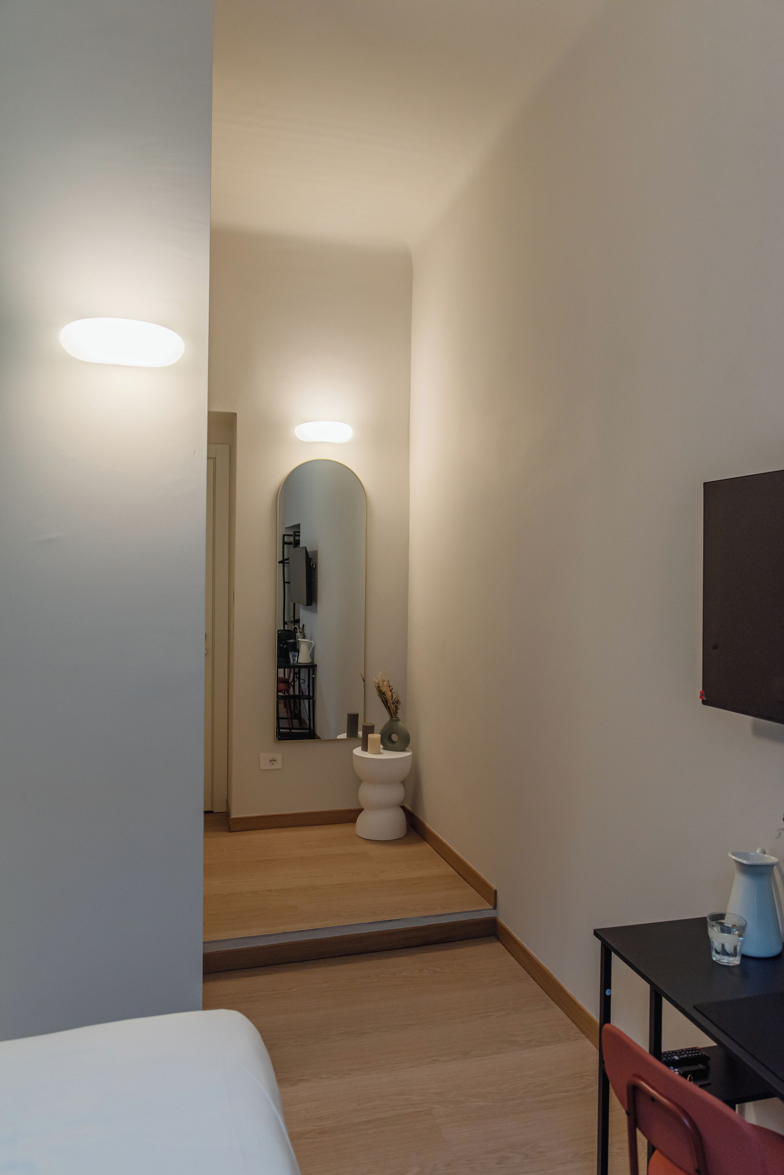 Un couloir étroit, avec un lit sur la gauche, une petite table et une chaise sur la droite, et des toilettes avec un miroir au centre.