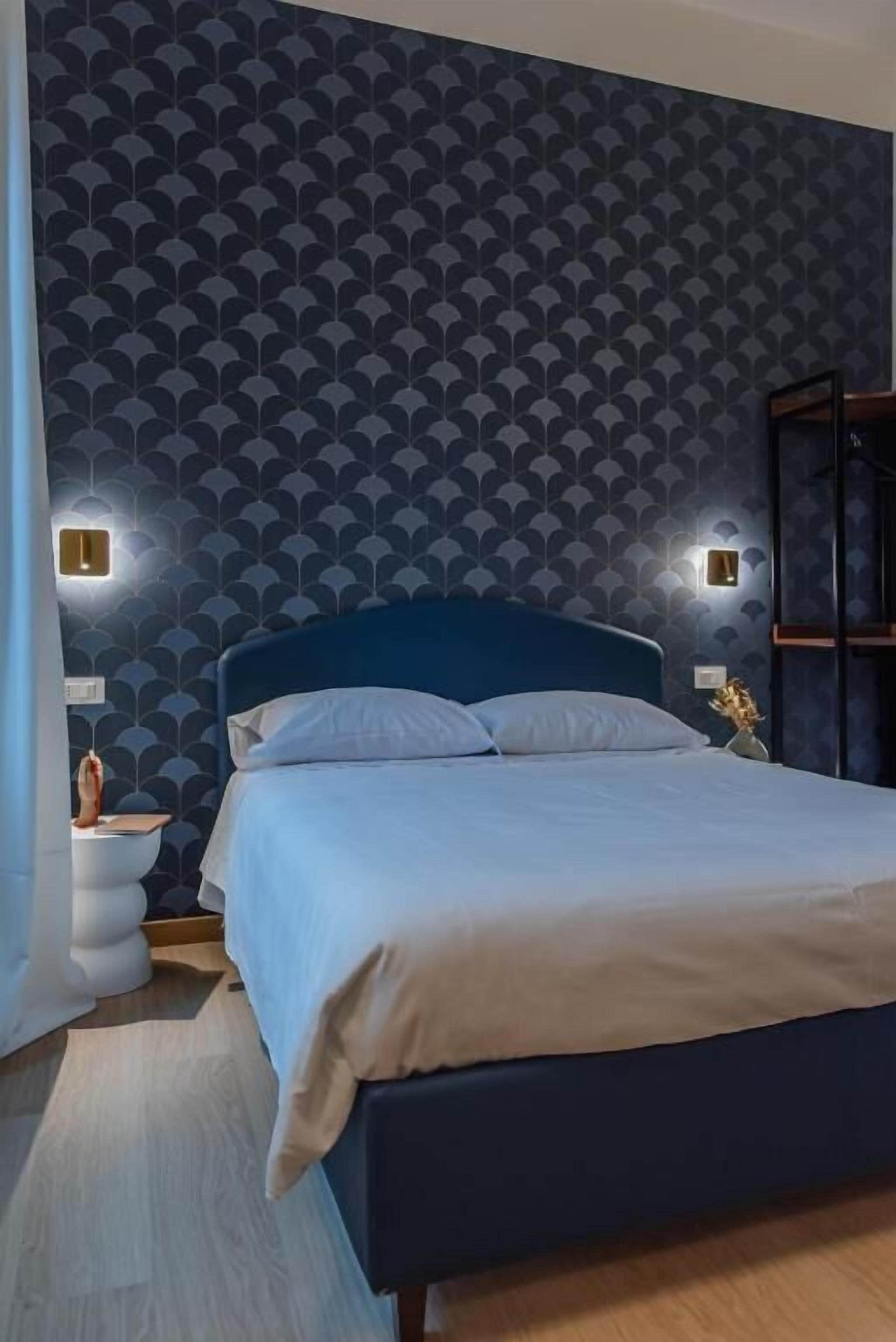 A bedroom with a dark blue headboard, a bed with white bedding, and a patterned wallpaper. There are two wall-mounted lights on either side of the bed.