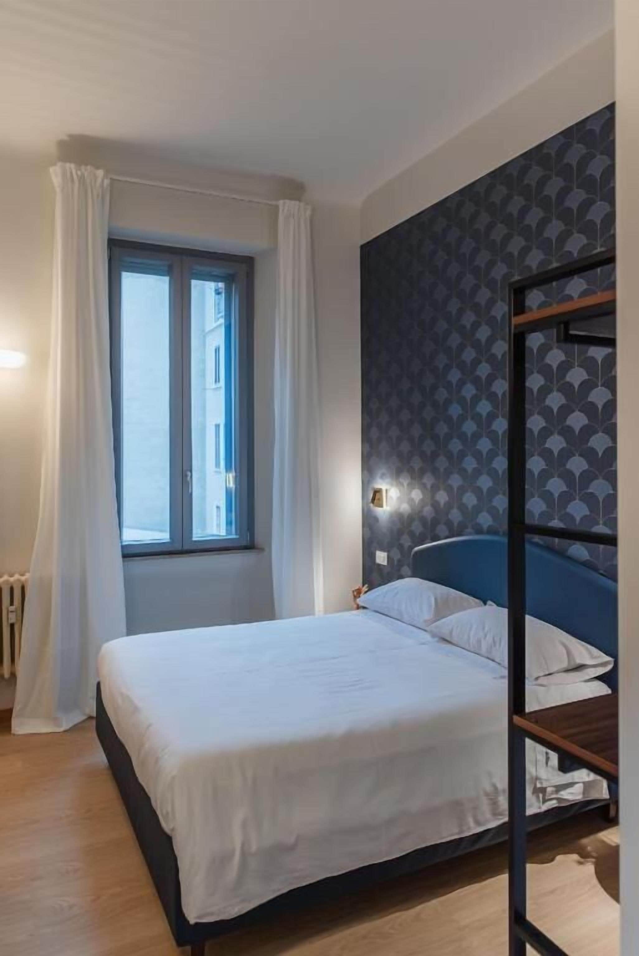 A bedroom with a bed, a window with curtains, a wall with a geometric pattern, and a radiator.