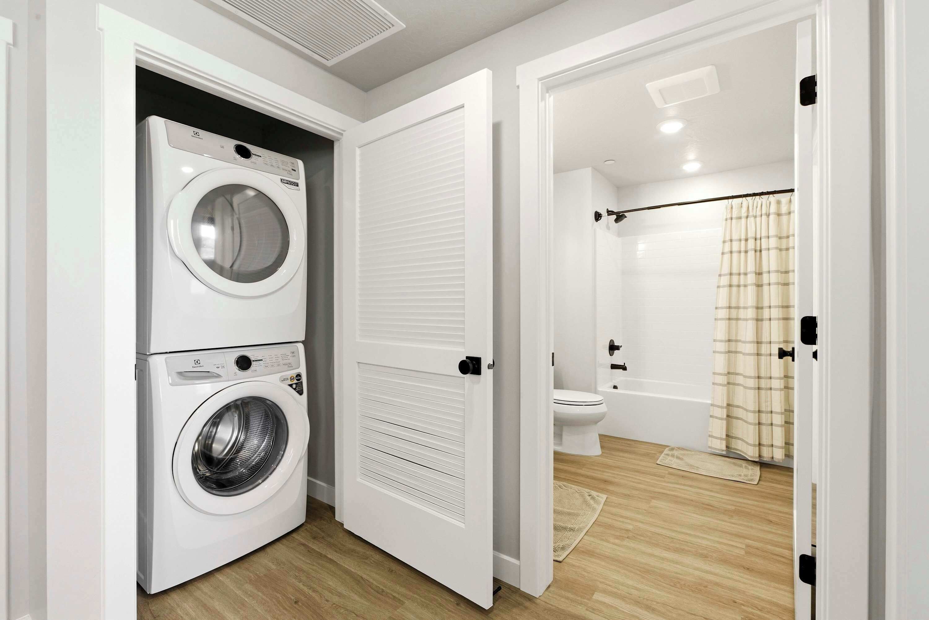 A laundry room with a front-loading washer and dryer, a toilet, and a shower area with a curtain.
