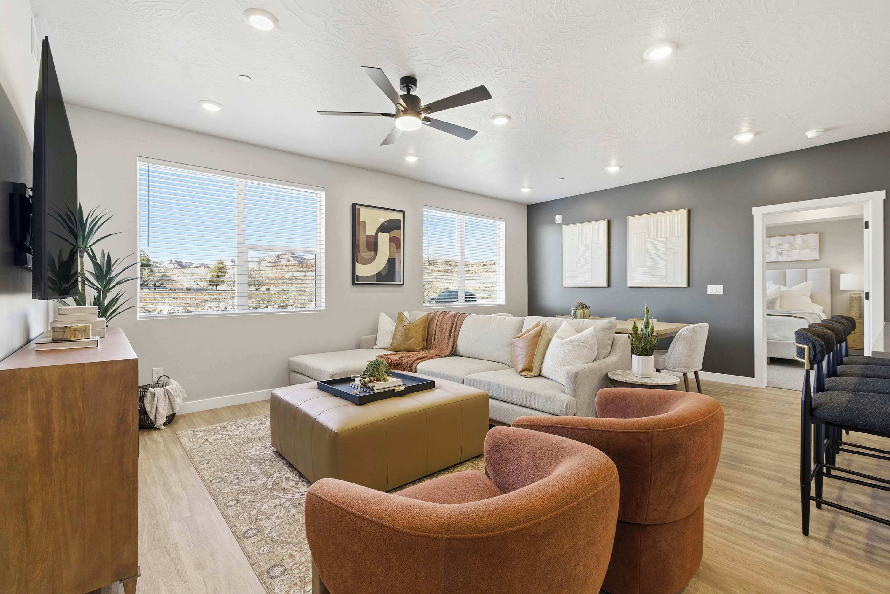 A modern living room with a sofa, ottoman, and chairs, a ceiling fan, and large windows.