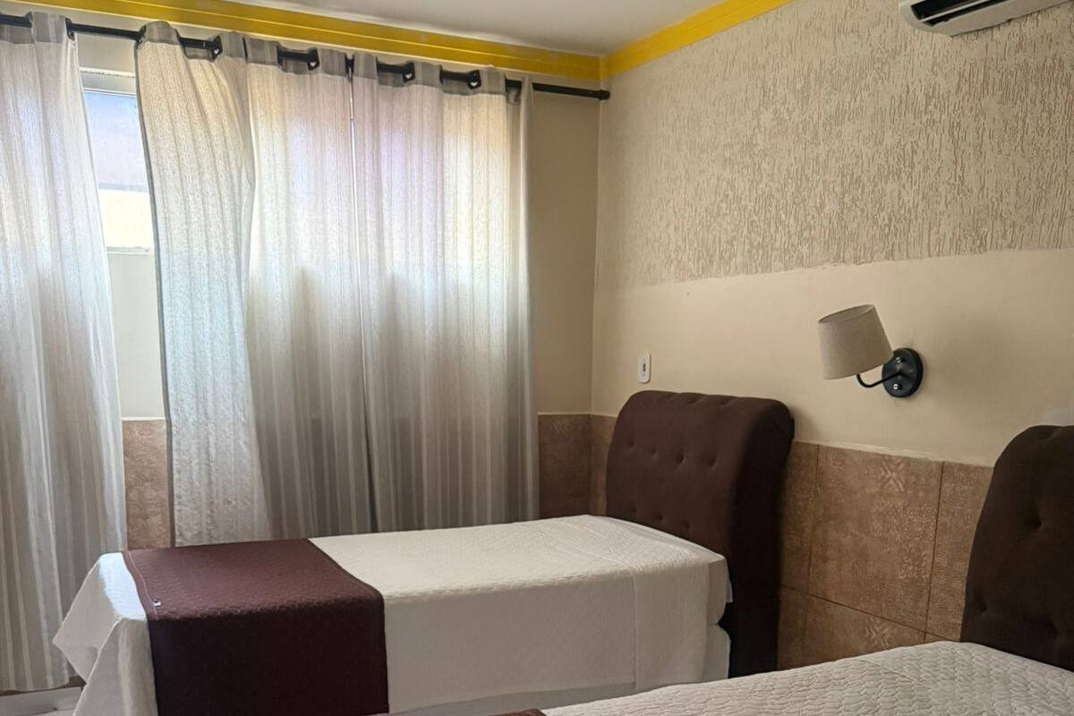 A hotel room with two single beds, a window with curtains, a wall-mounted air conditioning unit, and a wall lamp.