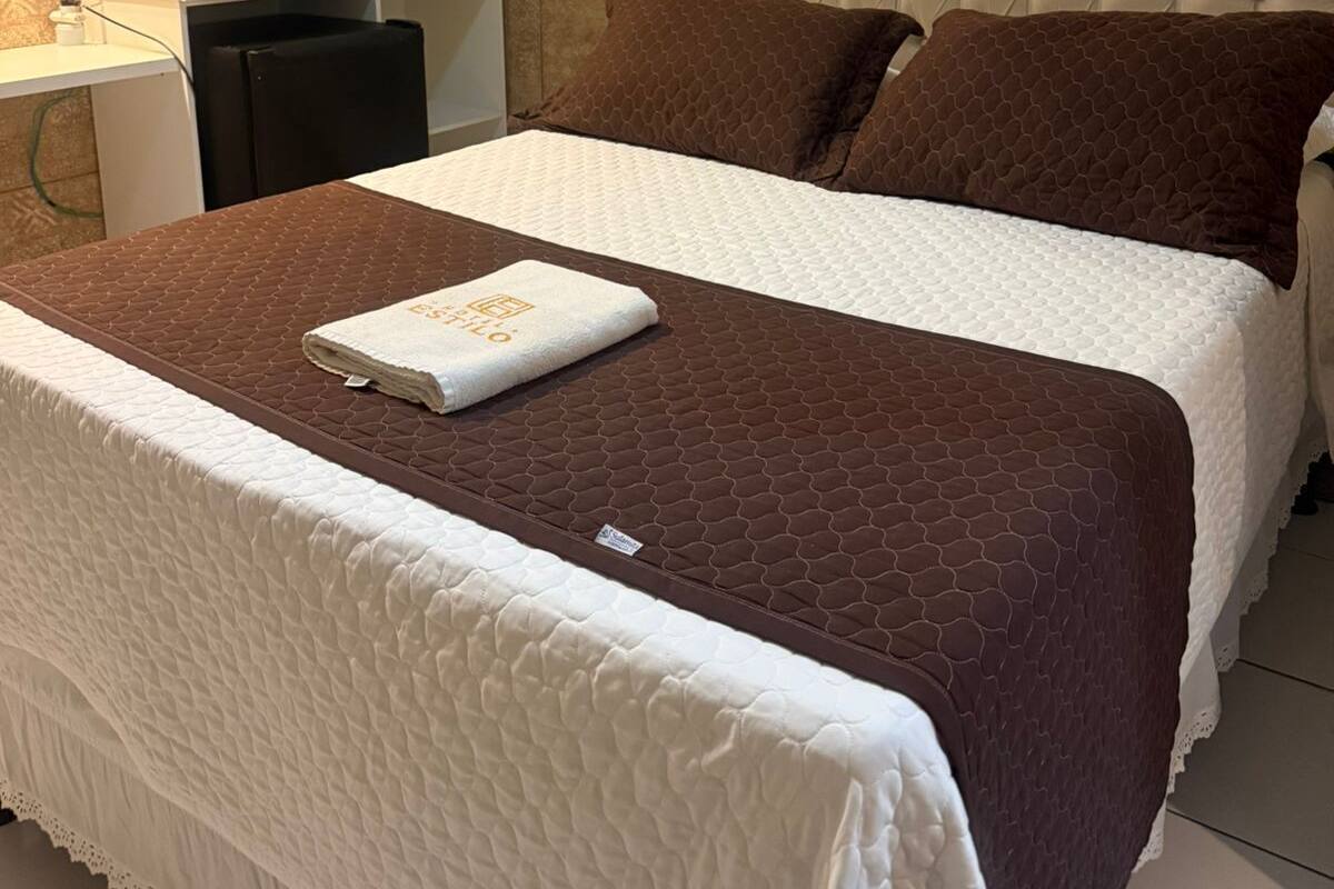 A neatly made bed with a brown quilt, a white duvet, and two brown pillows. There is a white wardrobe with a yellow accent on the wall behind the bed.