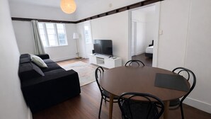 A modern living room with a black sofa, a wooden dining table with chairs, a flat-screen TV, and a large window with curtains. - Affordable house, walk to South Bank, CBD & transport (South Brisbane)