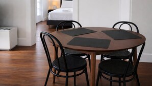 A round wooden table with four black chairs, a bed with white bedding, and a nightstand with a lamp in a room with white walls and wooden flooring. - Affordable house, walk to South Bank, CBD & transport (South Brisbane)