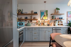 Une cuisine équipée d’un four à micro-ondes, d’un grille-pain et de divers ustensiles de cuisine rangés sur des étagères. On y trouve également une table à manger avec des chaises et un luminaire suspendu.
