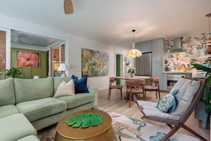 Un salon moderne avec un canapé vert, un coin repas et une cuisine décorée d’un papier peint aux motifs tropicaux.