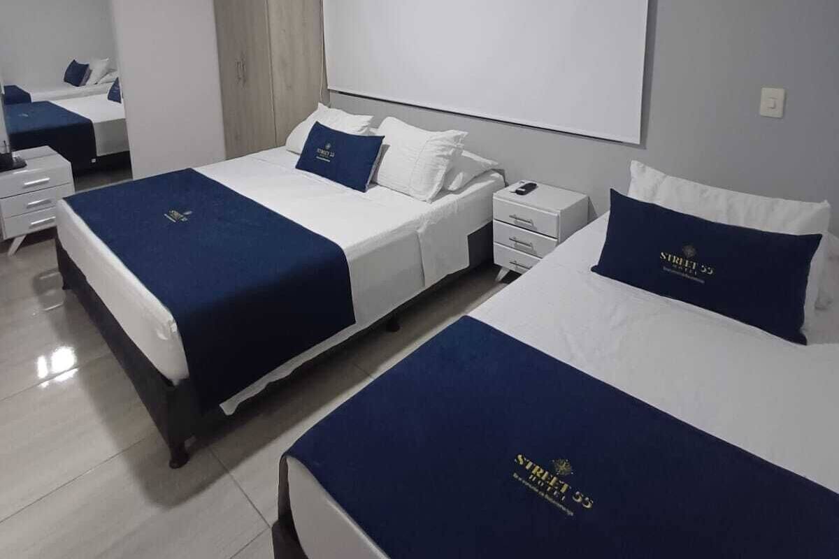 Ein Hotelzimmer mit zwei Betten, jedes mit einem blauen Kopfteil und weißer Bettwäsche. Ein großes Fenster bietet Blick auf den Himmel.