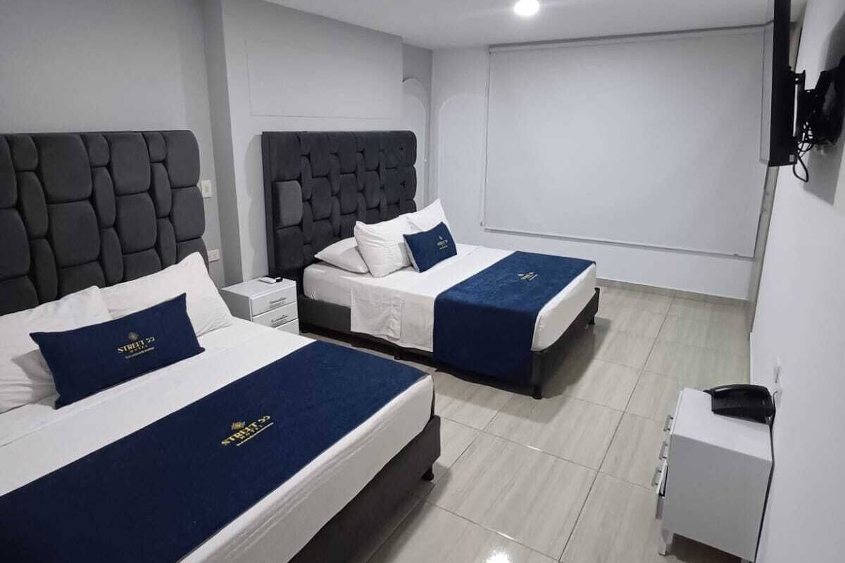 Ein Hotelzimmer mit zwei Betten, jeweils mit Kopfteil, weißer Bettwäsche und blauen Ziertelementen. Auf einem kleinen Nachttisch stehen eine Lampe und ein Telefon.