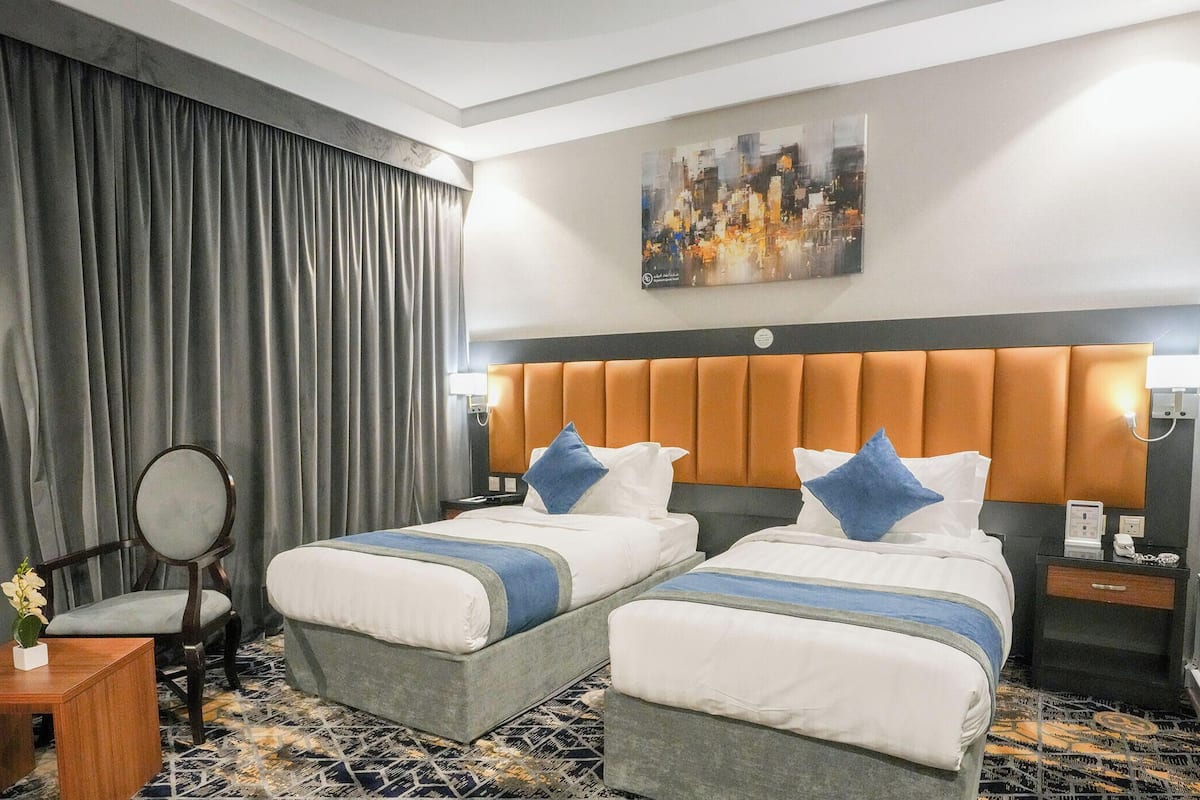 Habitación de hotel con dos camas, una mesita de noche, una silla, un escritorio, un cuadro en la pared y un ventanal con cortinas.