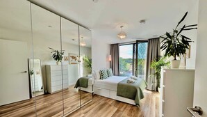 A modern bedroom with a large mirror, wooden flooring, a bed with green bedding, and a view of greenery outside. - Charming Flat Near London Attractions (Sheerwater)
