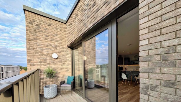 A modern balcony with a wooden floor, a seating area with a chair and a small table, and a glass door leading to an interior space with a kitchen visible. - Charming Flat Near London Attractions (Sheerwater)