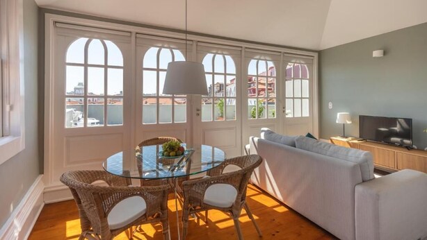 A living room with a glass table, wicker chairs, a grey sofa, and a television on a wooden cabinet. - Clube Charming Apartments - Ribeira (Porto)