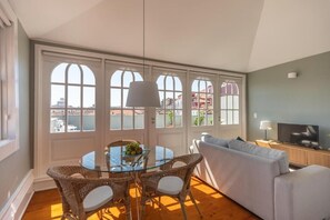 A living room with a glass table, wicker chairs, a grey sofa, and a television on a wooden cabinet. - Clube Charming Apartments - Ribeira (Porto)