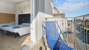 A balcony with a blue safety rail, a bed with a geometric-patterned bedspread, and a sliding door leading to a room with a wooden panel wall. - Apartment 'Villa Trentina Trilocale 2 Piano' with Shared Terrace and Wi-Fi (Jesolo)