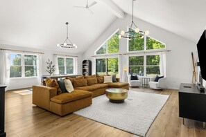 A spacious living room with a large sectional sofa, a coffee table, and a fireplace. There are multiple windows providing natural light and views of the outdoors. - Innsbruck Chalet | Family-Friendly | Game Room | Fire Pit | Hot Tub (Boyne City)