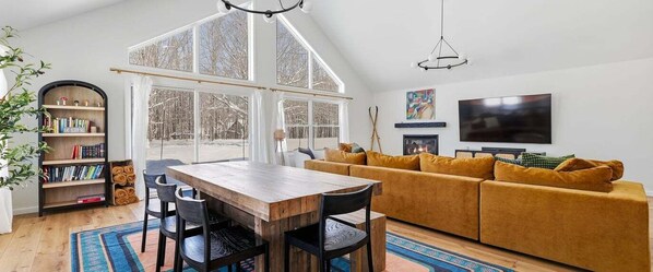 A cozy living room with a wooden dining table, a sofa, a fireplace, and a TV. - Innsbruck Chalet | Family-Friendly | Game Room | Fire Pit | Hot Tub (Boyne City)