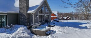 A house with a chimney, a hot tub, and a snow-covered yard. - Innsbruck Chalet | Family-Friendly | Game Room | Fire Pit | Hot Tub (Boyne City)