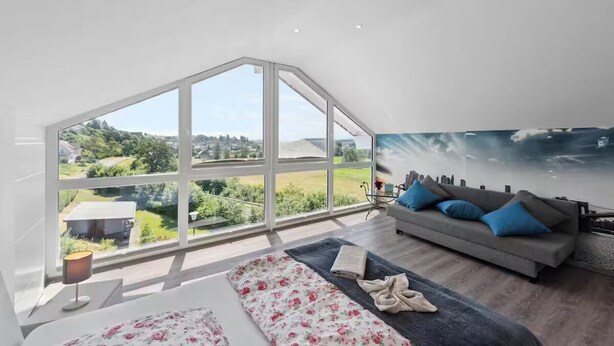 Et moderne soveværelse med et stort ovenlys, en sofa med blå puder og udsigt til grøn natur udenfor.