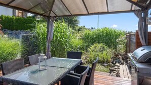 En udendørs terrasse med et glasbord, fletstole, grill og et overdækket område med udsigt til grøn bevoksning og en bygning.
