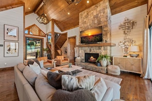 A spacious living room with a stone fireplace, a large TV mounted above it, and a comfortable seating area with a sofa and chairs. - No Cabin Pressure - Mountain Views | Hot Tub | Outdoor Fireplace (Blue Ridge)