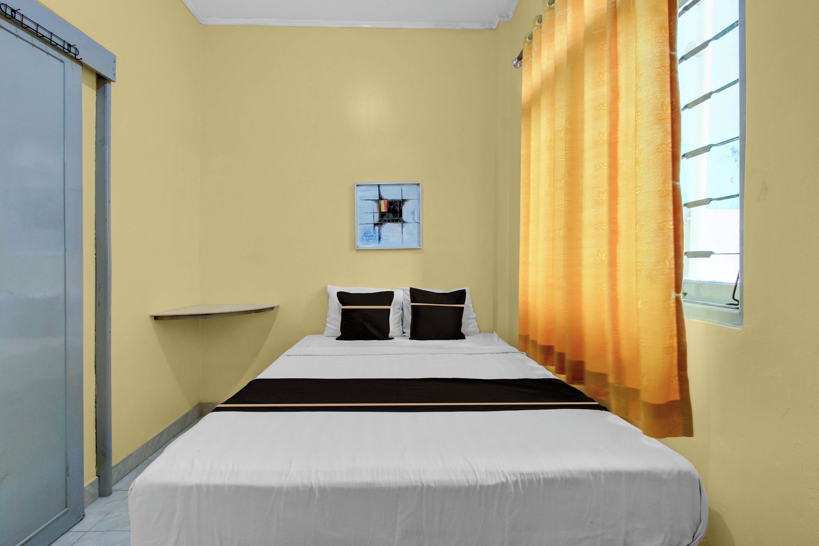 A hotel room with a bed, a window with curtains, a wall-mounted artwork, and a door.