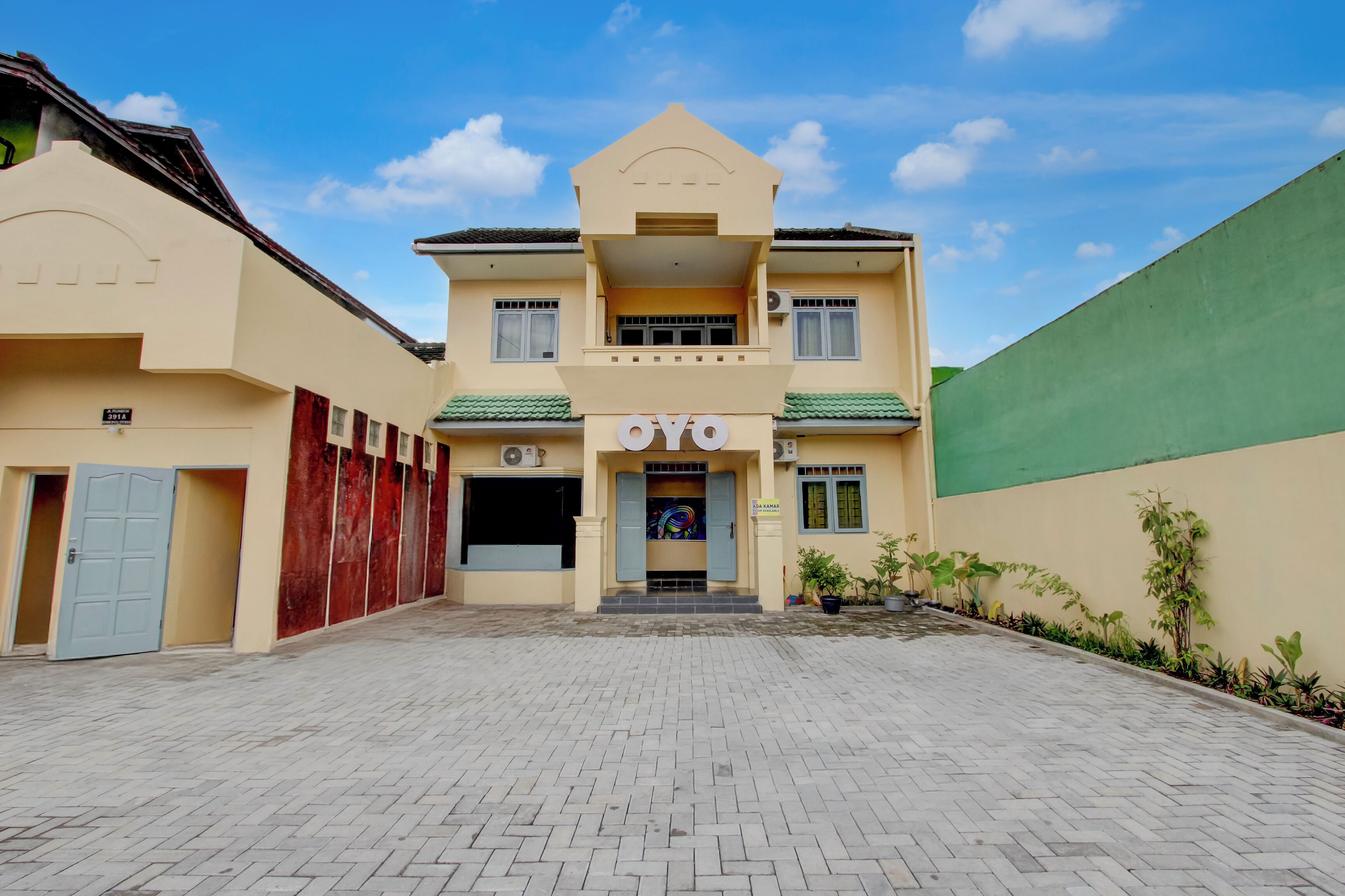 Un edificio de dos pisos con un cartel que dice 'OYO' en la entrada, un patio empedrado y un muro verde a la derecha.