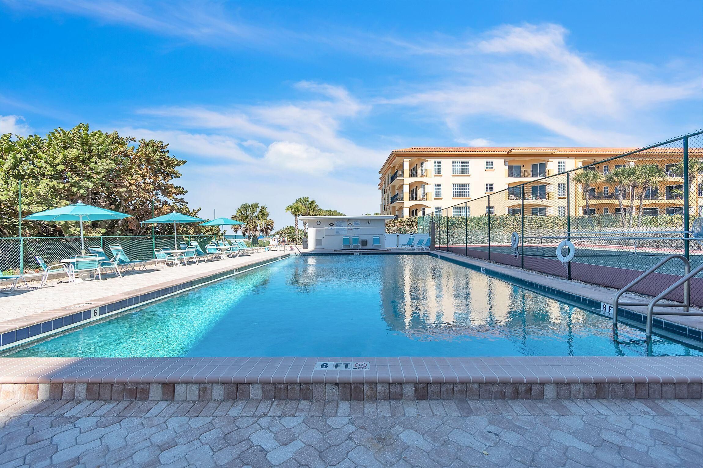 A swimming pool with lounge chairs and umbrellas, surrounded by a fence and a building in the background.
