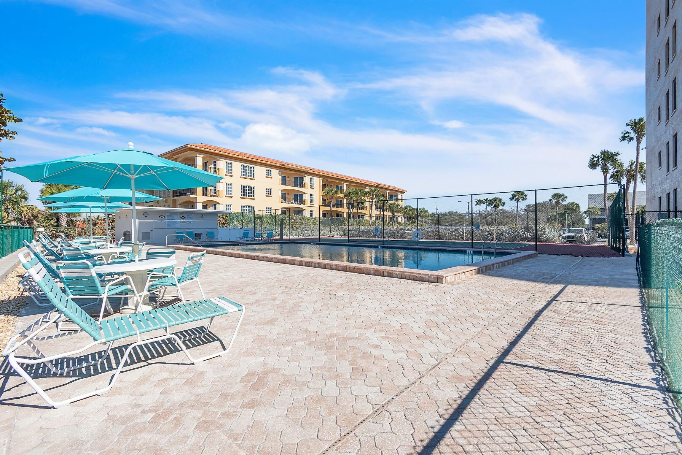 Un área de piscina con sillones de descanso y sombrillas, una piscina y un edificio con balcones al fondo.
