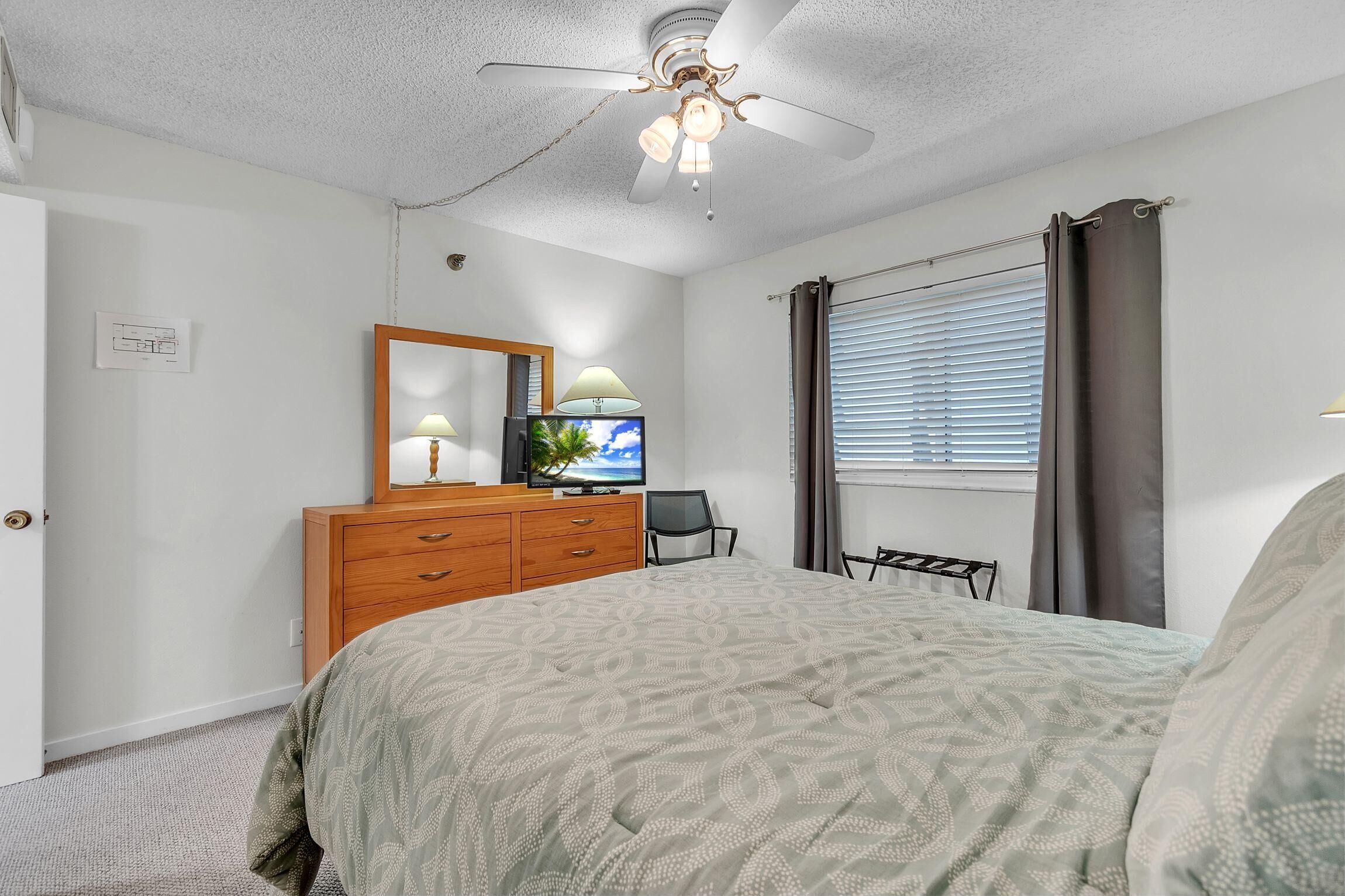 Ein Schlafzimmer mit Bett, Kommode, Fernseher, Deckenventilator und Fenster mit Vorhängen.