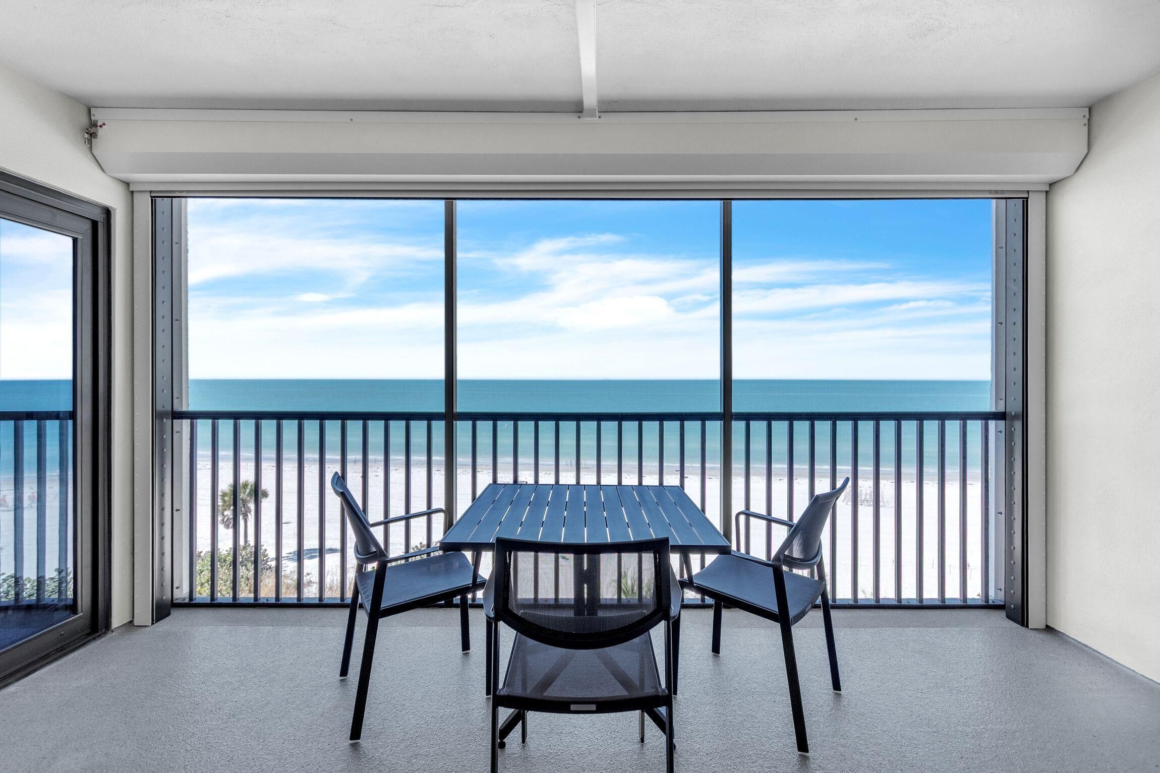Un balcon avec une table et des chaises, donnant sur une plage sous un ciel dégagé.