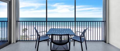 Un balcon avec une table et des chaises, donnant sur une plage sous un ciel dégagé.