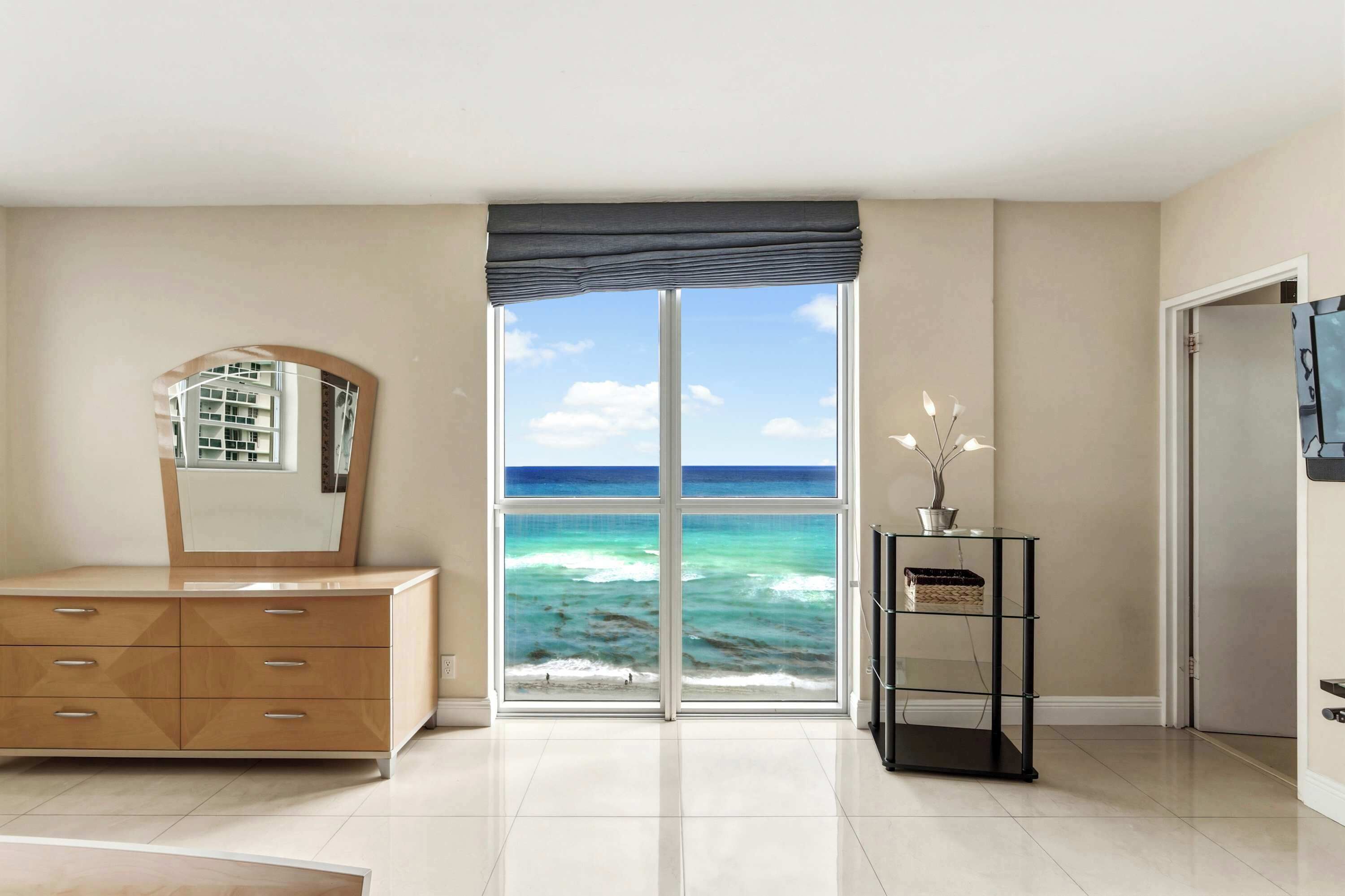A room with a large window, a dresser, a mirror, a small table with a vase, and a door.