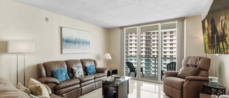 A modern living room with a leather sofa, a glass coffee table, a brown armchair, and a large window with a view of high-rise buildings.