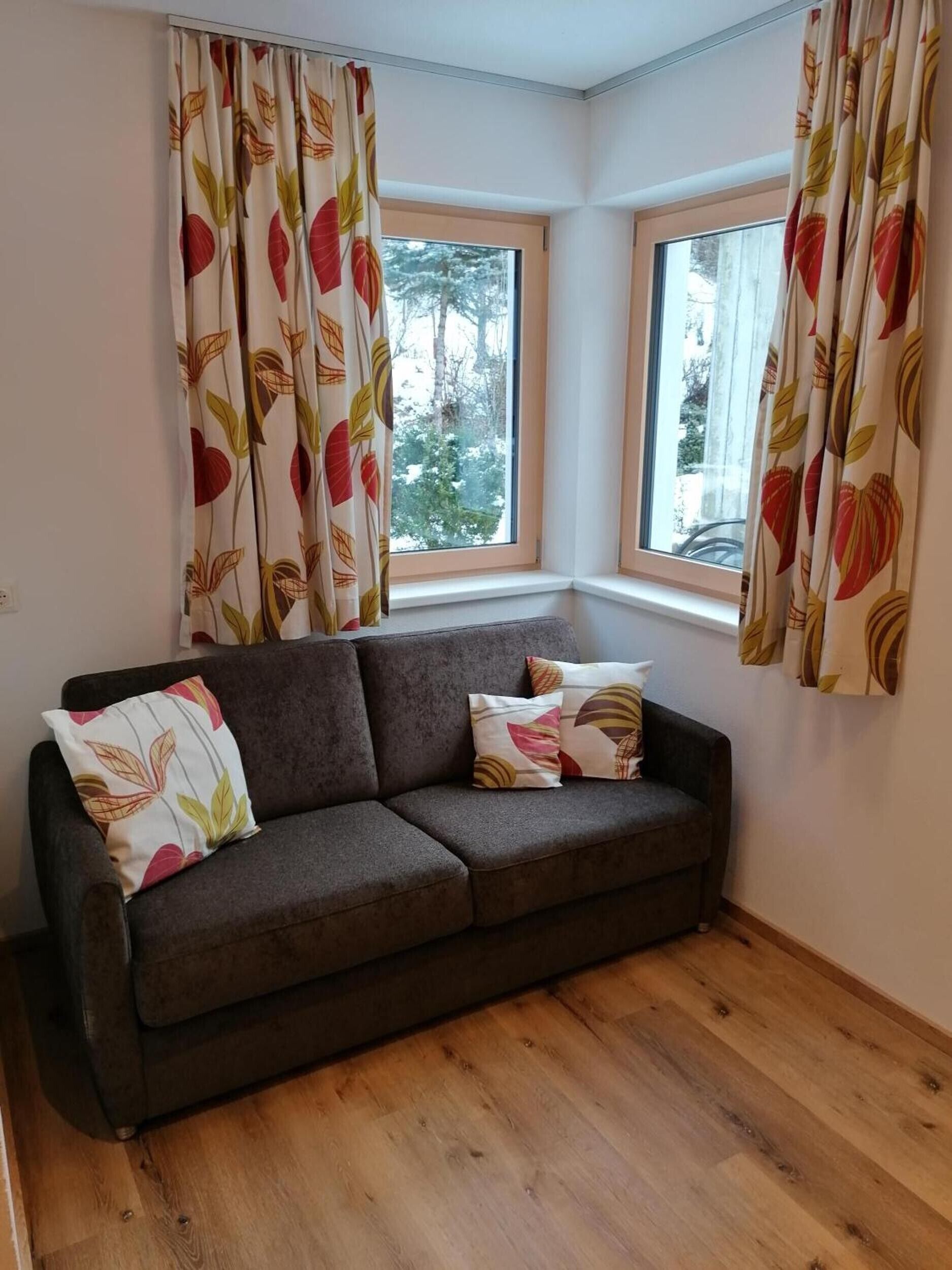 Habitación de esquina con un sofá gris oscuro, dos almohadas y una ventana con cortinas.