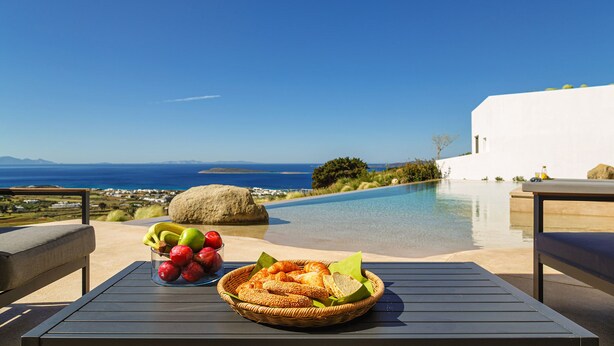 Mesa com tigela de frutas e cesta de pão, com vista para a piscina e oceano.