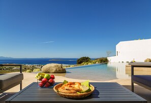 Et bord med frugtskål og brødkurv, med udsigt til pool og hav.