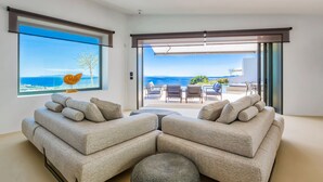 Uma sala de estar moderna com uma grande janela que oferece vista para o mar, um sofá modular, um banquinho redondo e uma área de pátio com mobiliário para área externa.