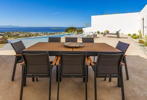 An outdoor dining area with a wooden table and chairs, overlooking a pool and a scenic view of the ocean and landscape. - Paros Villa Skylines Estate II (Paros)